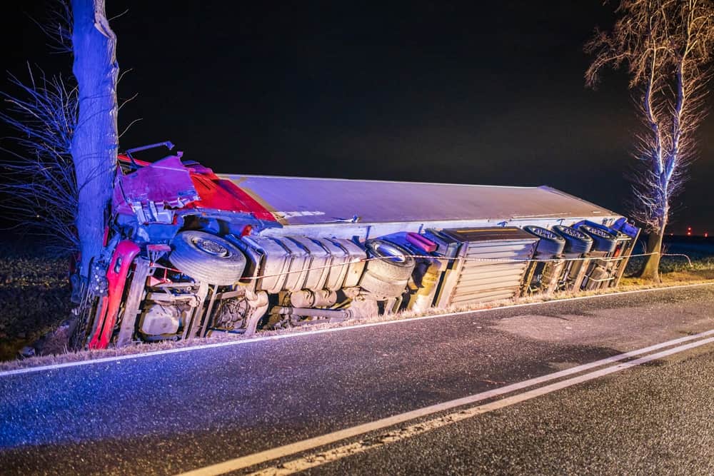 A semi-truck overturned on the side of the road at night near a tree, with emergency lights illuminating the scene
